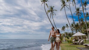 A couple walking along a concrete path beside the ocean, with palm trees and lounge chairs in the background. - Kubu Indah Dive & Spa Resort (Kubu)