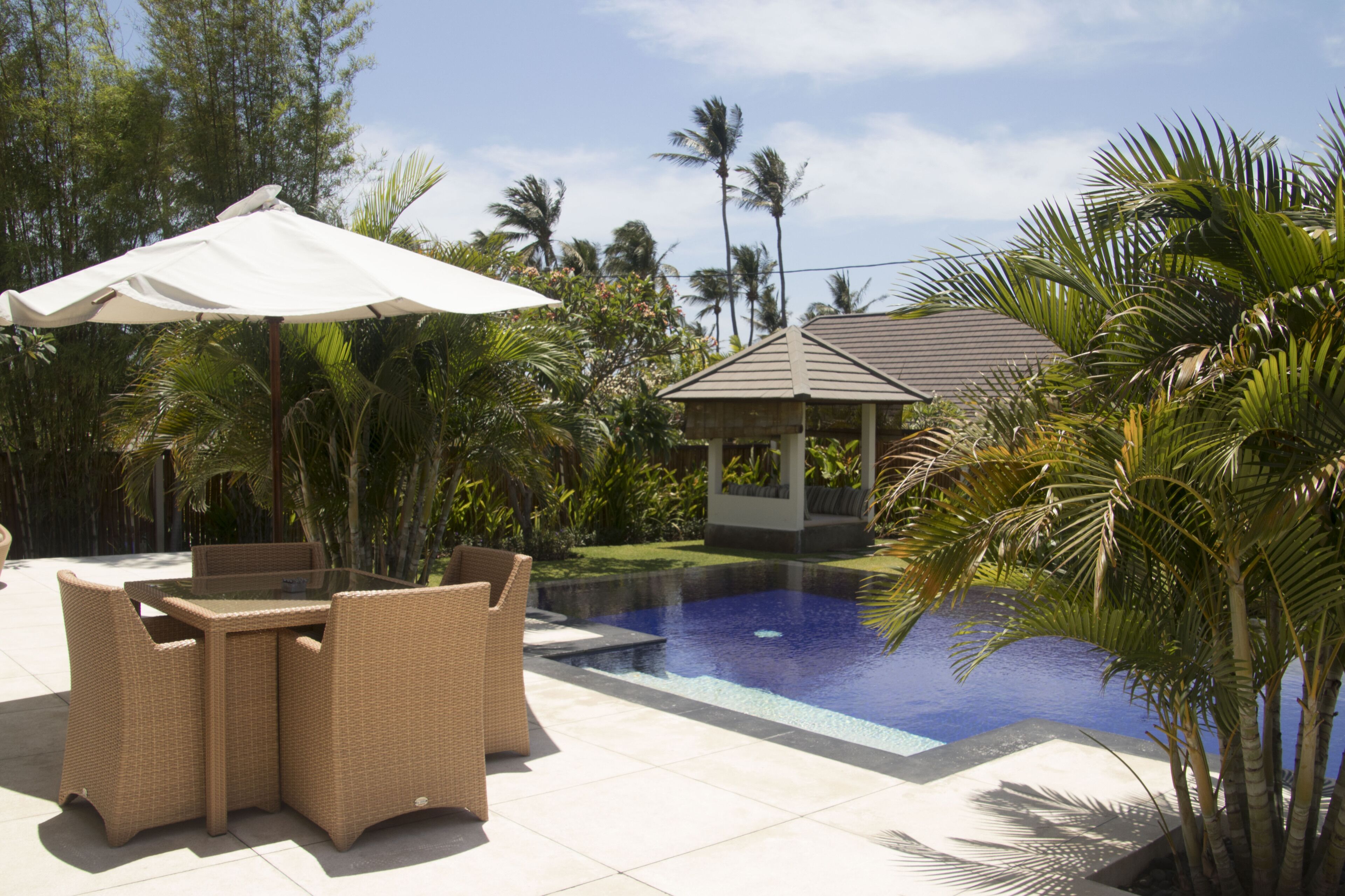 A pool area with a glass table and chairs, surrounded by palm trees and a gazebo.