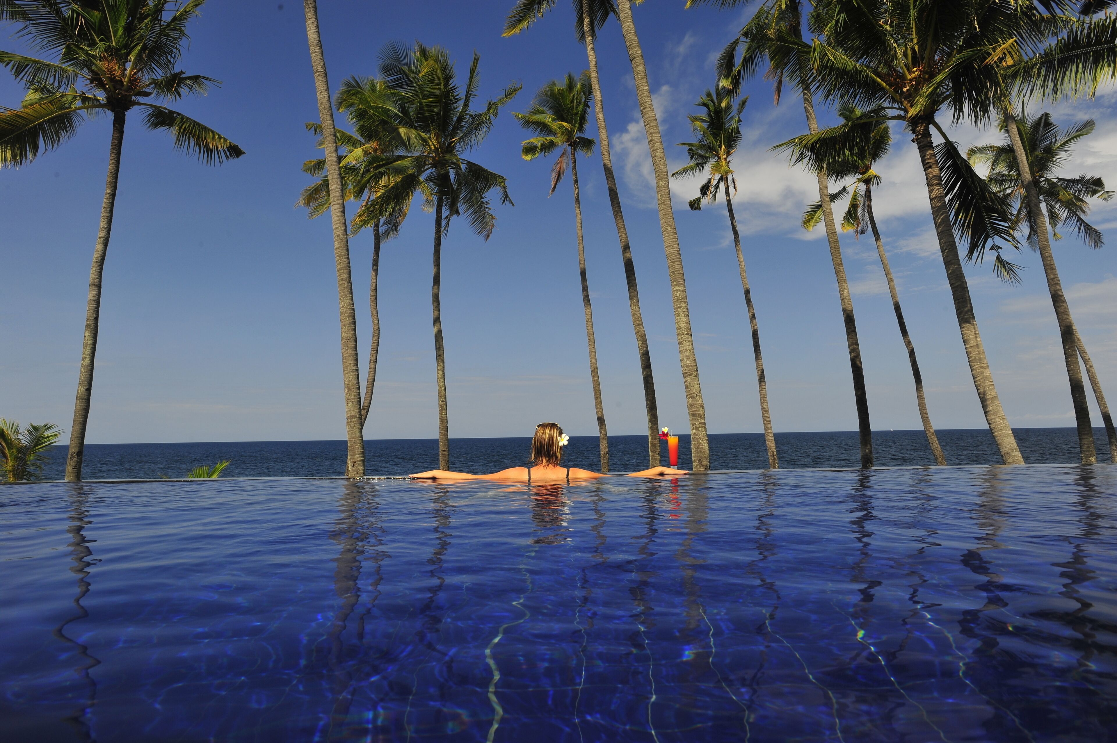 Una persona relajándose en una piscina infinita de color azul cristalino, rodeada de altas palmeras, con vistas al océano.