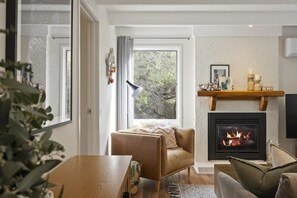 Ein gemütliches Wohnzimmer mit Kamin, einer bequemen Couch, einem Esstisch und Blick ins Freie durch ein großes Fenster.