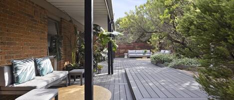 A wooden deck with a sofa and a bench, surrounded by greenery and a brick wall.