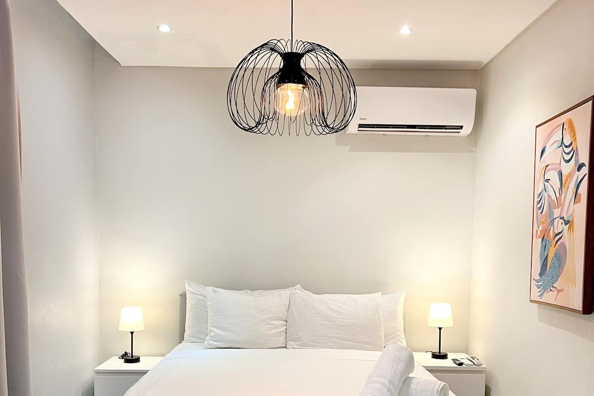 Une chambre d’hôtel moderne dotée d’un grand lit, de deux tables de chevet avec lampes, d’un luminaire au plafond original et d’un climatiseur fixé au mur.
