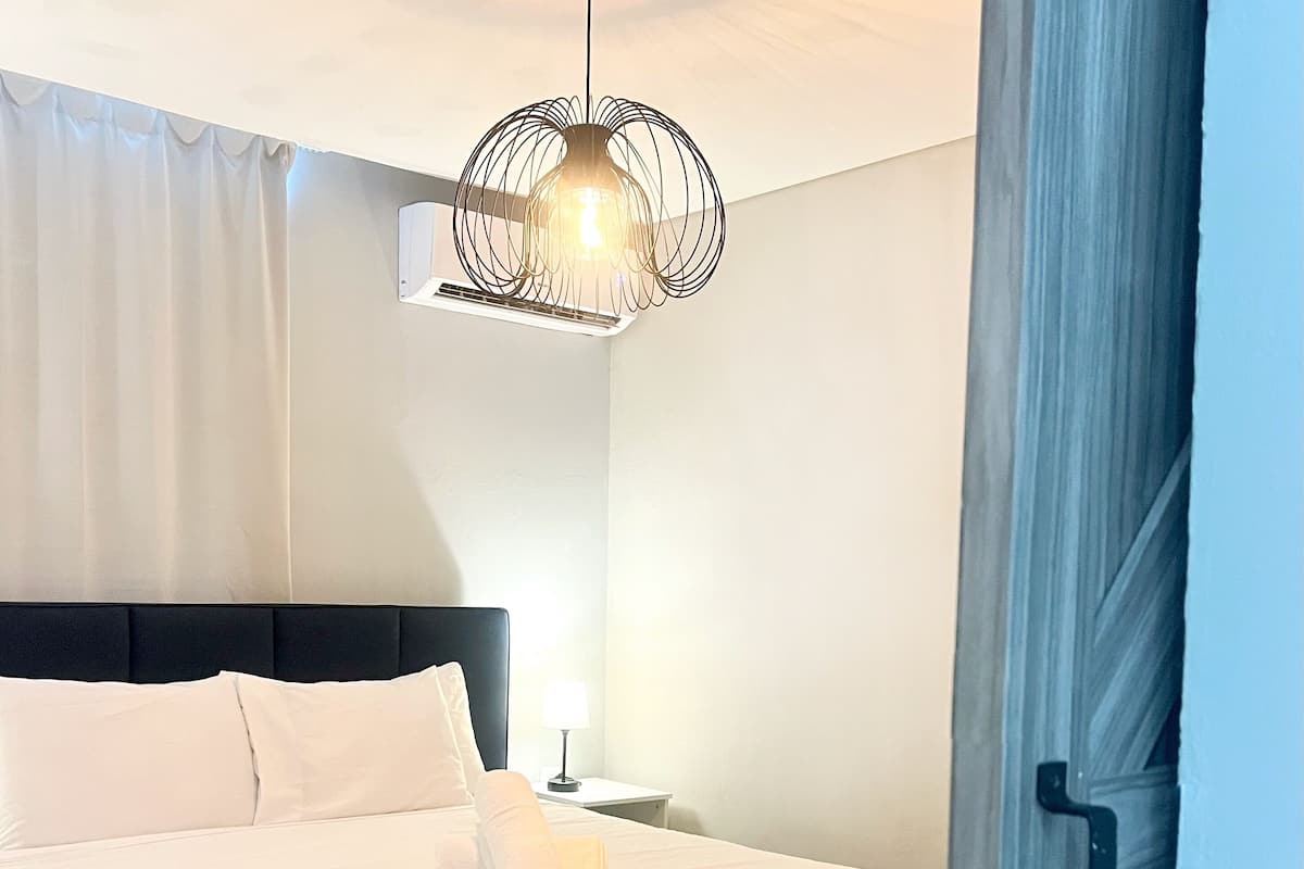 Un lit bien fait, avec du linge de lit blanc et des oreillers, un luminaire suspendu moderne et un plancher en bois.