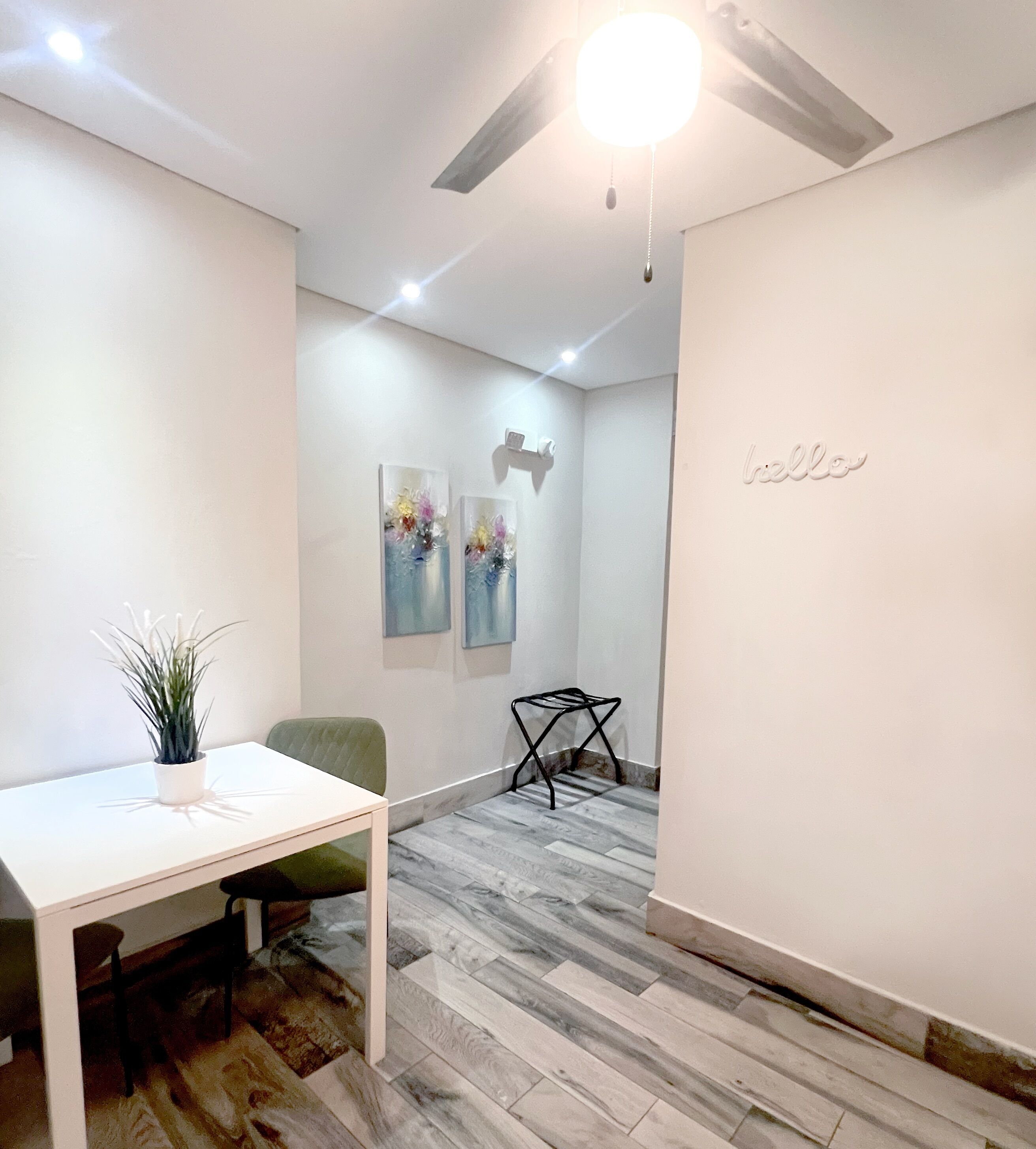 A modern, minimalist room with a white table, a green chair, and a ceiling fan. There are two abstract paintings on the wall and a 'hello' sign.