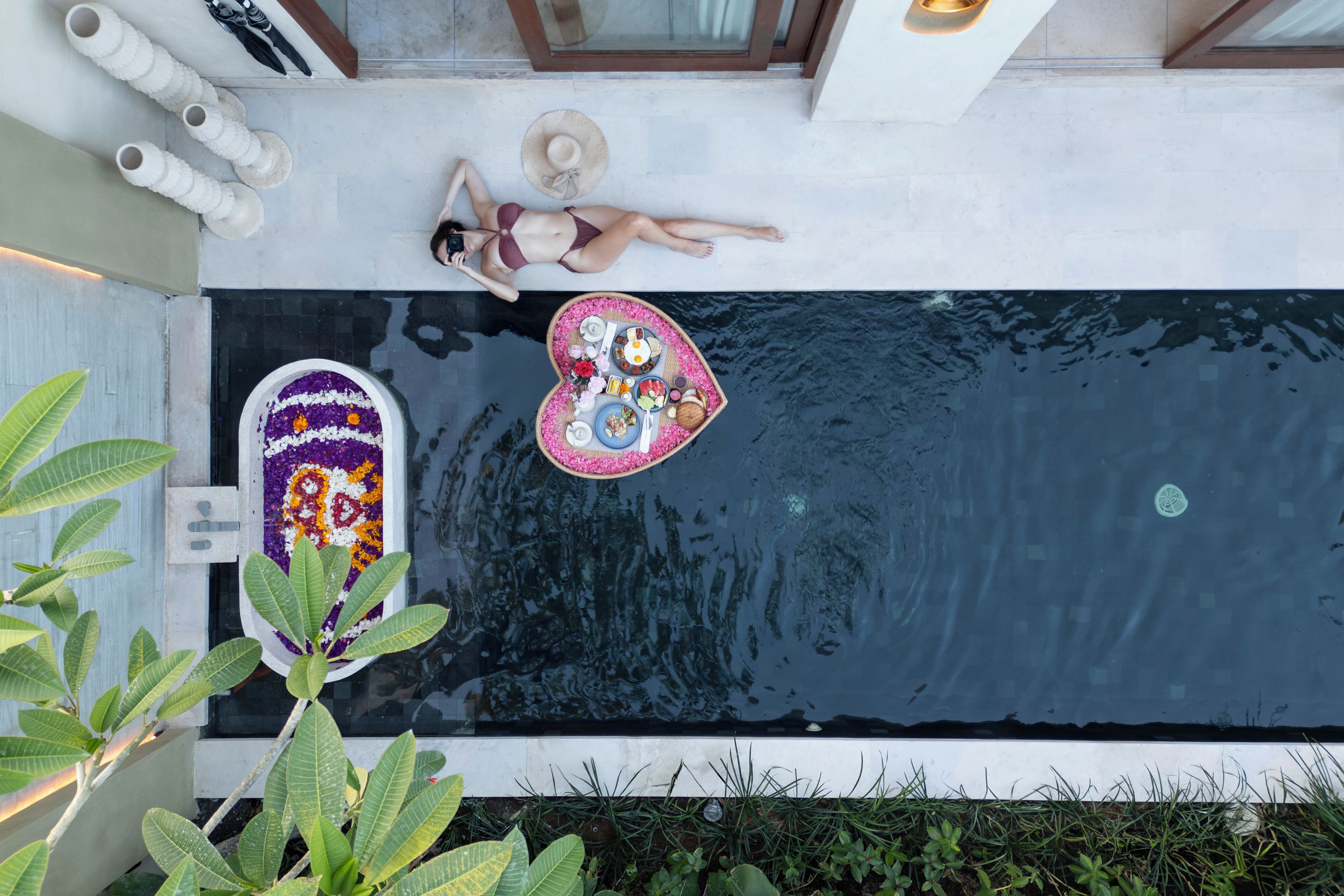 A pool with a floating platform, a woman lying on the edge, and a decorative arrangement of flowers and fruits.