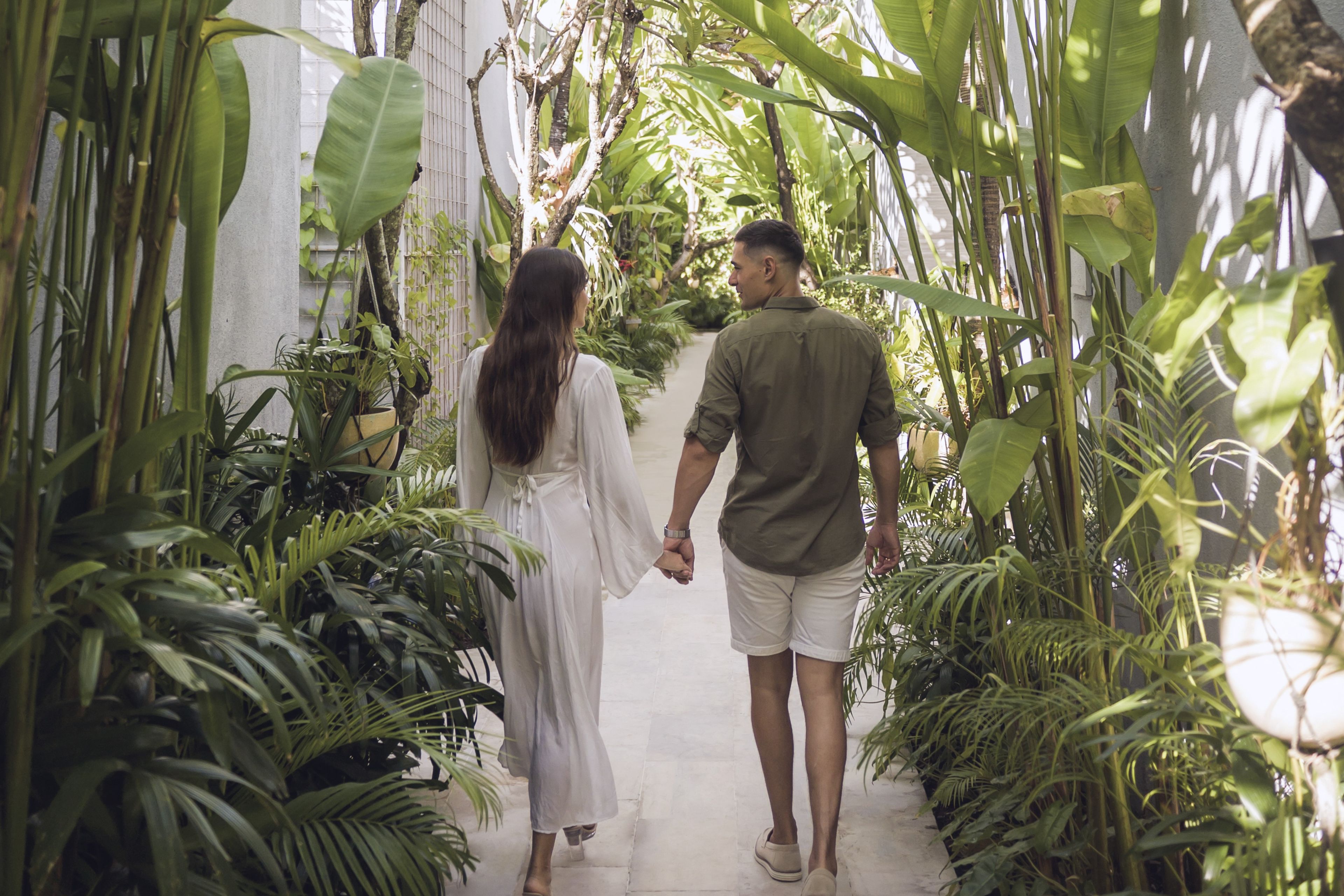 A couple walking hand in hand down a narrow pathway surrounded by lush greenery and plants.