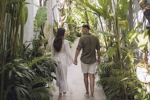 A couple walking hand in hand down a narrow pathway surrounded by lush greenery and plants.