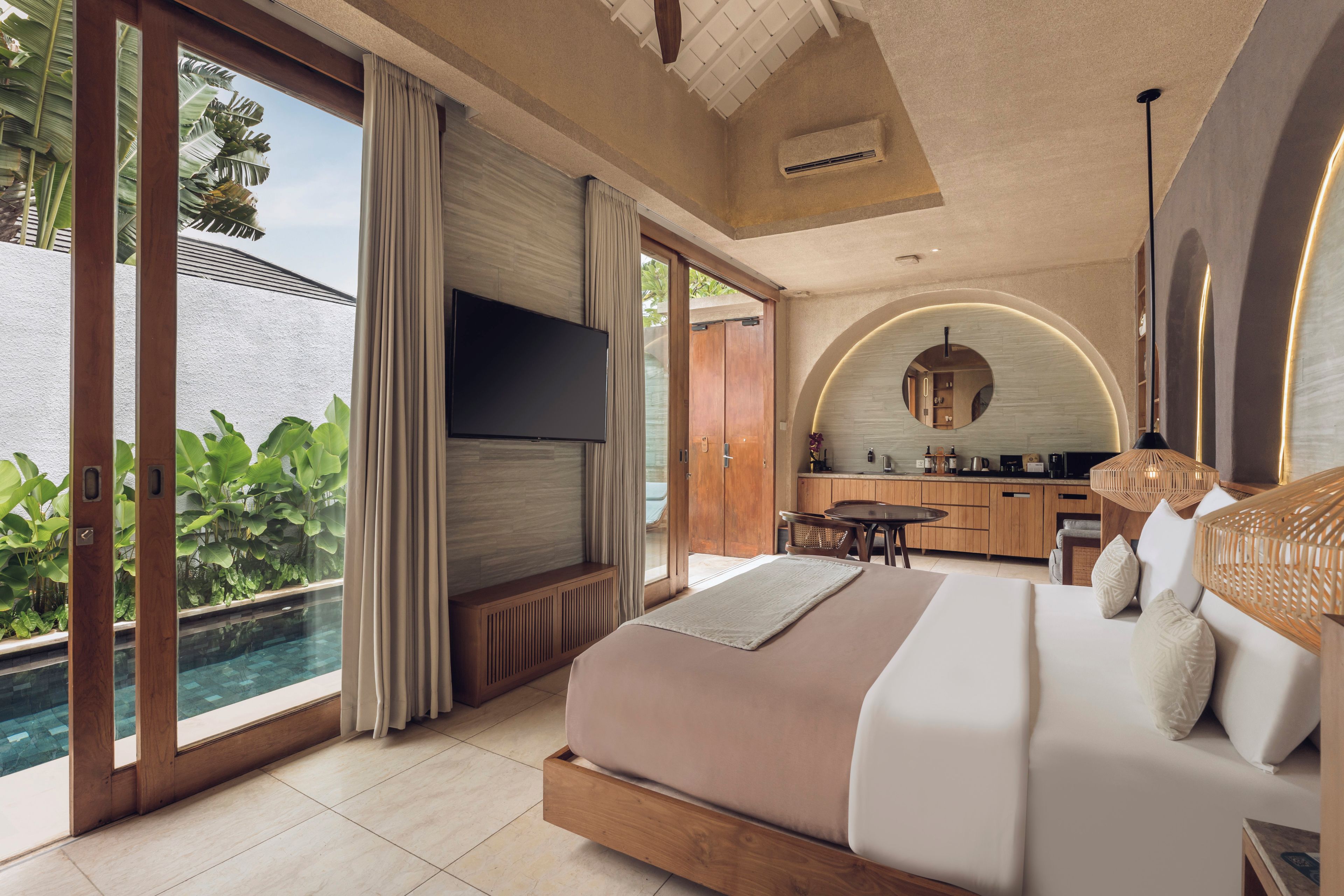 A modern bedroom with a large bed, a TV, and a view of a pool and greenery.