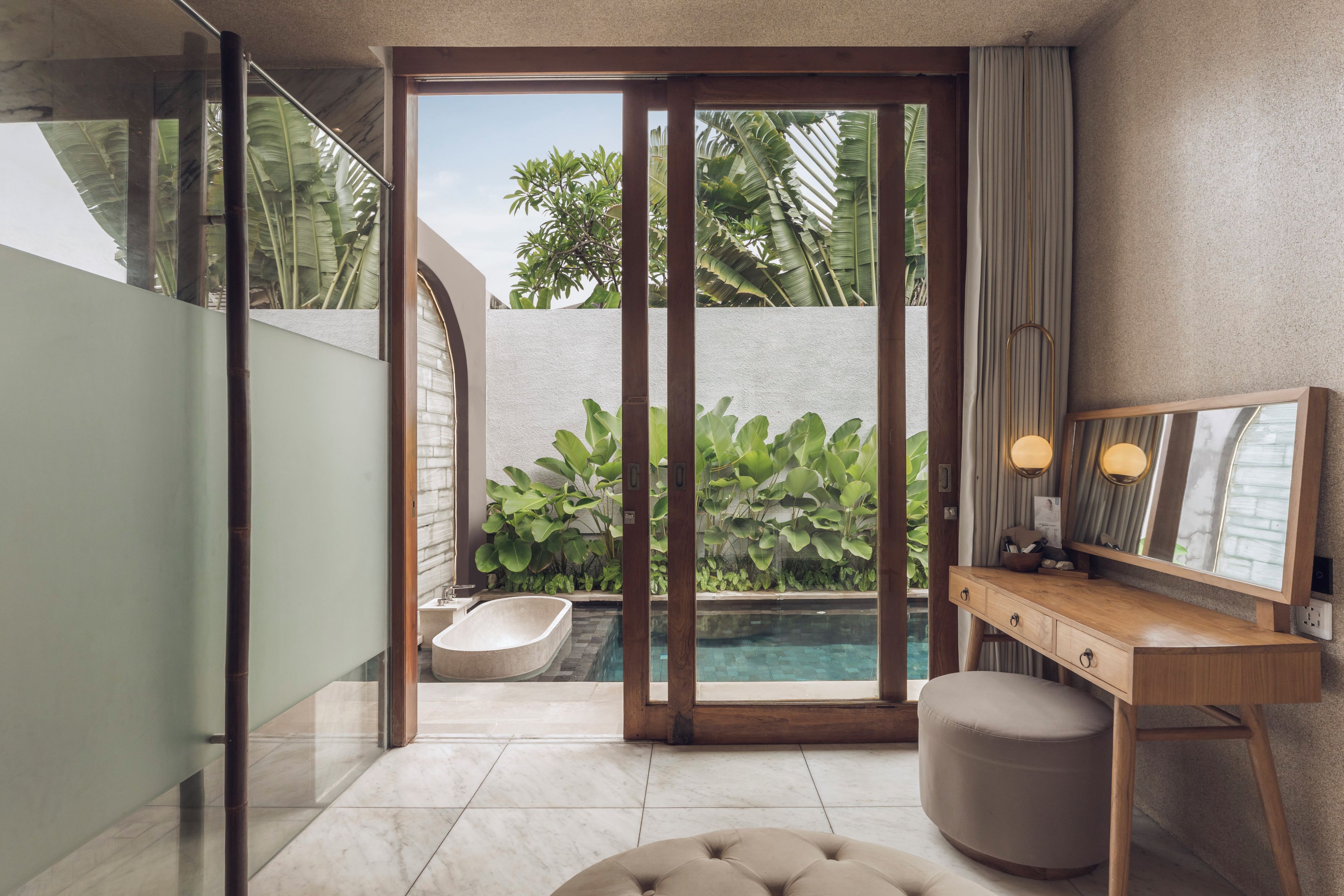 Un baño moderno con una puerta de vidrio que da a una piscina, un tocador de madera y suelo de mármol.