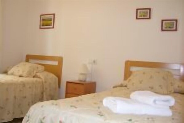 A room with two single beds, each with a wooden headboard and a floral-patterned bedspread. There is a small wooden nightstand with a lamp and a picture frame on the wall above the beds.