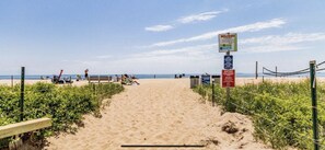 A sandy beach path leading to the ocean with a 'Welcome' sign, a warning sign, and a rope barrier.