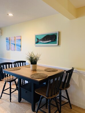 A dining area with a wooden table, black chairs, woven placemats, and wall art including a whale painting.