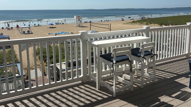 Vista de la playa desde una terraza de madera con barandas blancas, una torre de salvavidas y sillas de playa.