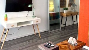 Een moderne woonkamer met een flatscreen-televisie, een bureau met een lamp, een houten salontafel en een spiegel die de kamer weerspiegelt.