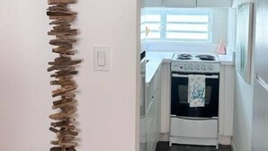 Een smalle gang met een verticale houten sculptuur links en een keuken met witte kasten en een raam rechts.