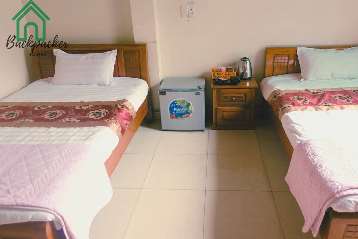 A hostel room with two beds, a small refrigerator, a nightstand, and a wall socket.