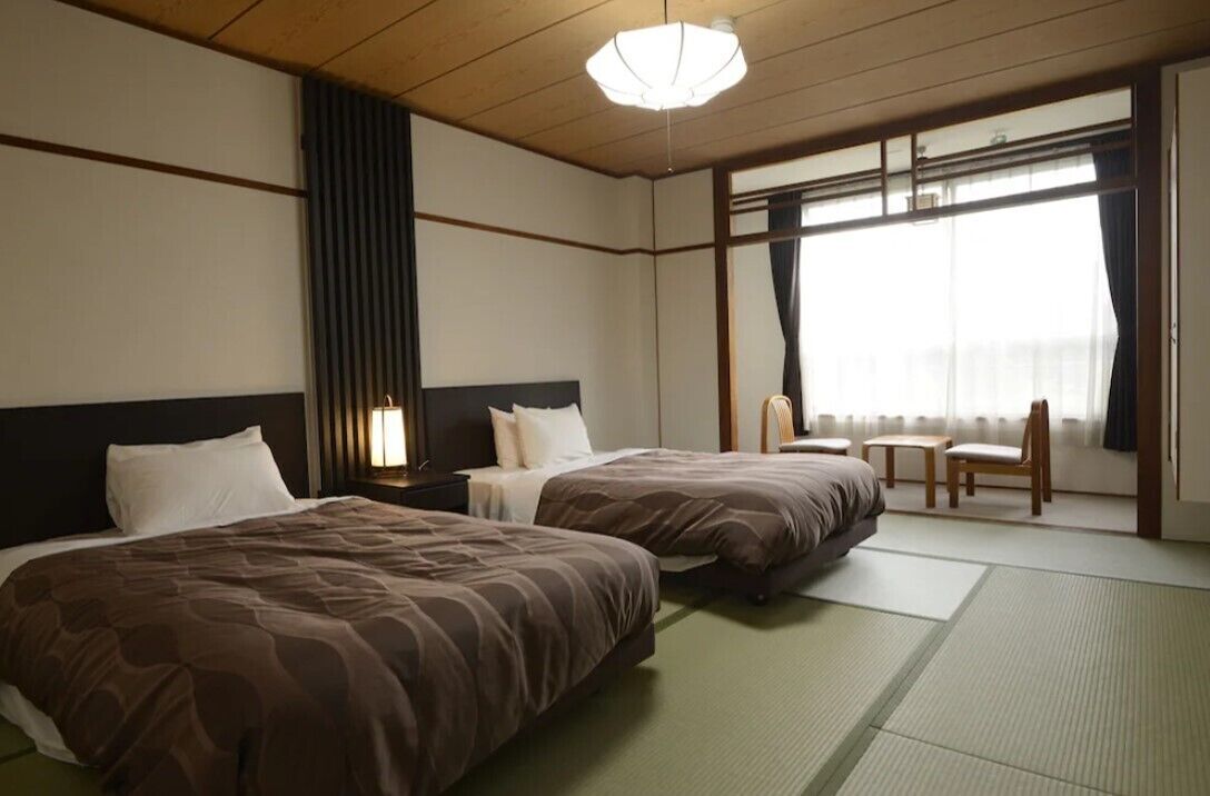 A hotel room with two beds, a nightstand, a chair, and a large window with curtains.