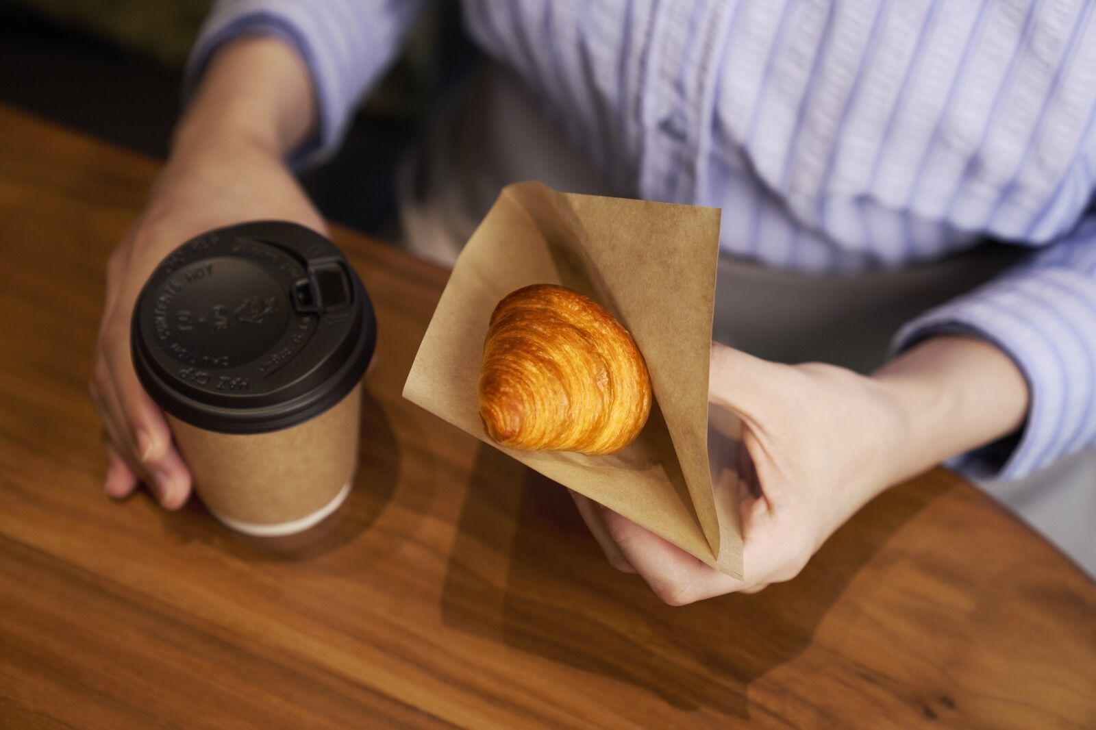 A person holding a paper bag with a croissant and a coffee cup.