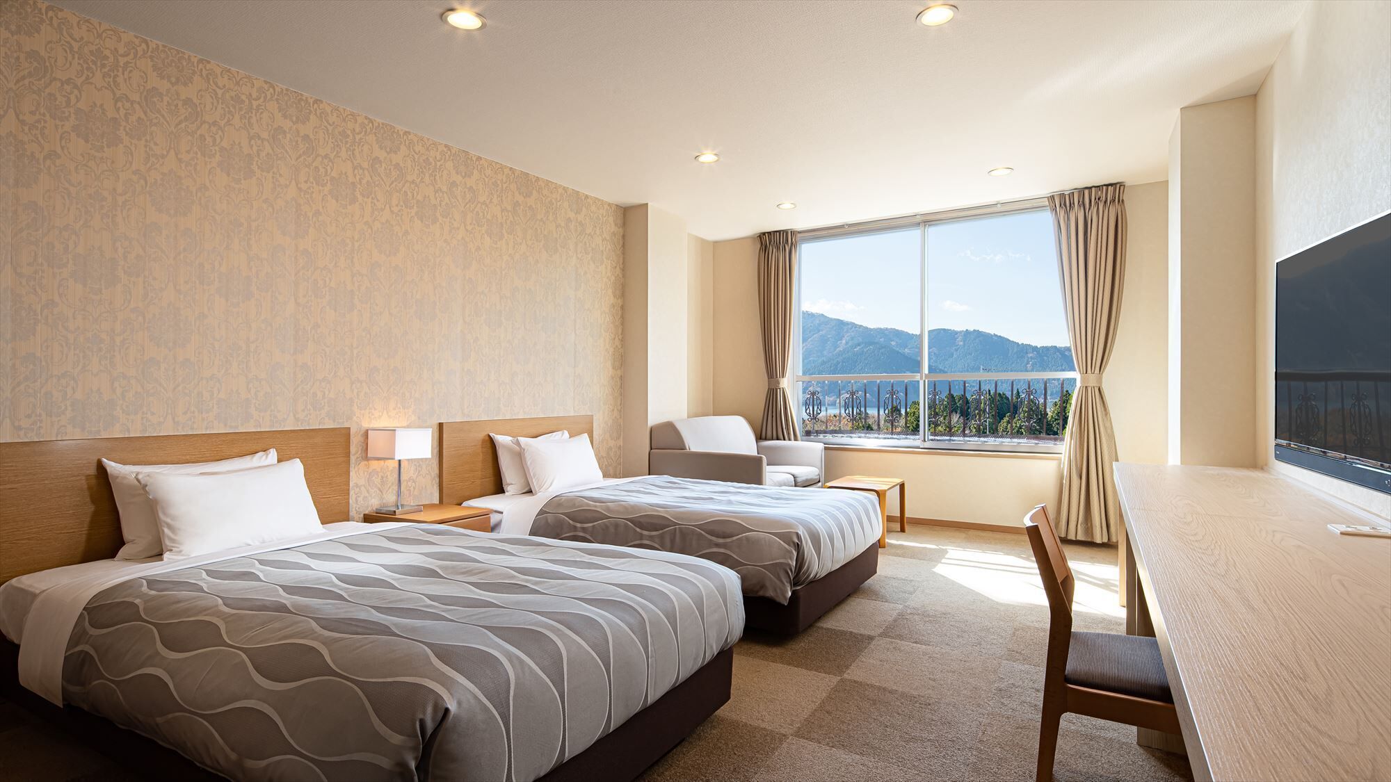 A hotel room with two beds, a desk, a chair, and a large window with a view of mountains.