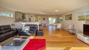 A spacious living room with a large black leather sofa, a glass coffee table, and a red rug. There is a dining area with a white table and chairs, and a kitchen area with cabinets and a sink.