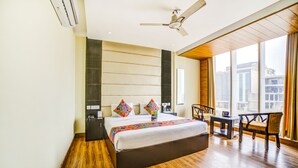 A modern hotel room with a large bed, a ceiling fan, a small table, and a view of the city through large windows. - FabHotel Diamond (Dera Bassi)