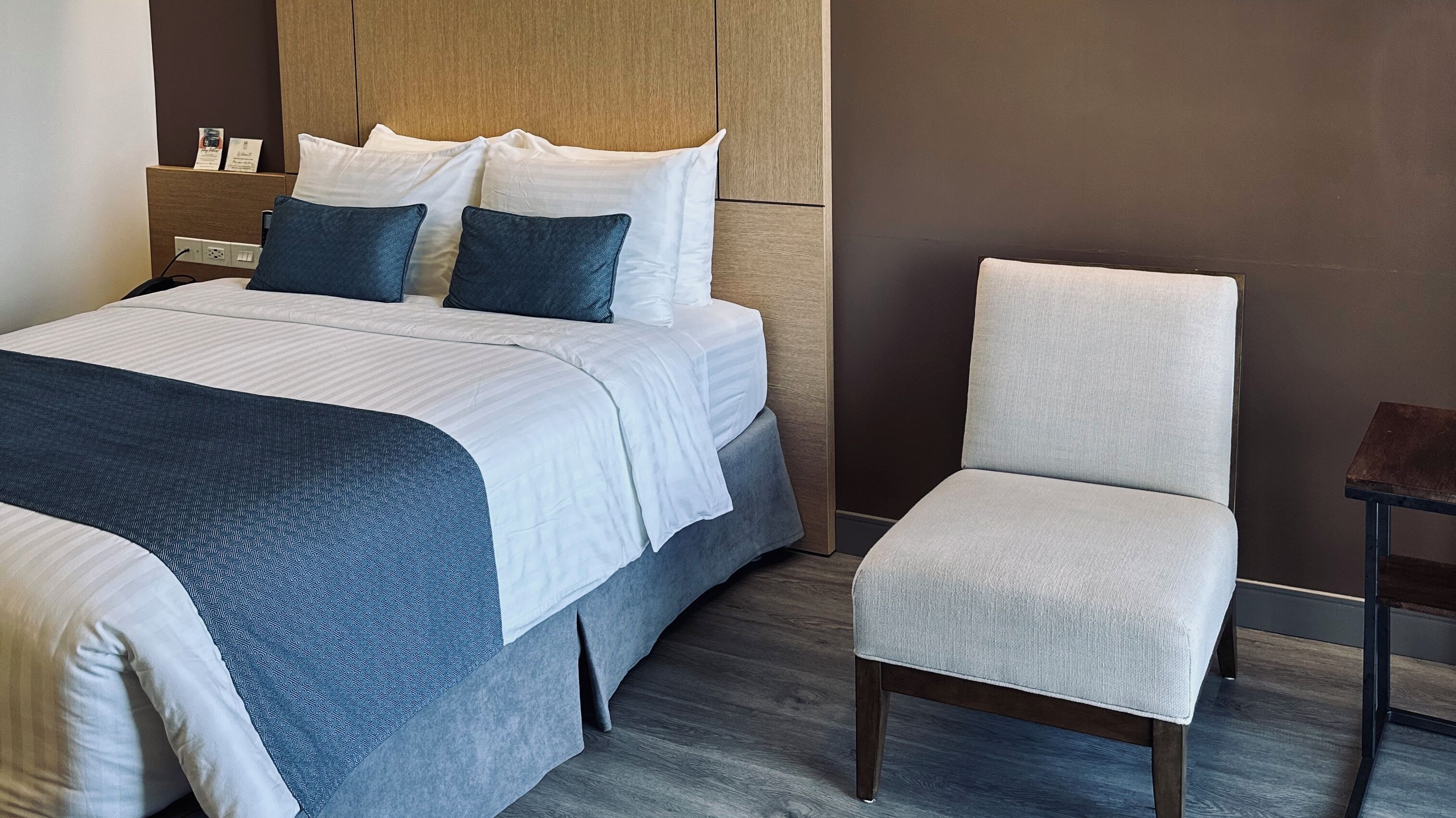 Habitación de hotel con cama, dos almohadas, mesita de noche y una silla.