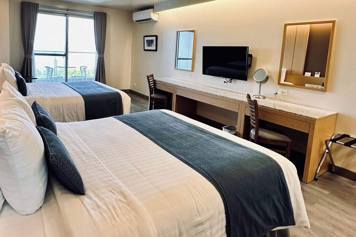 Habitación de hotel con dos camas, un escritorio, un televisor y un ventanal grande.