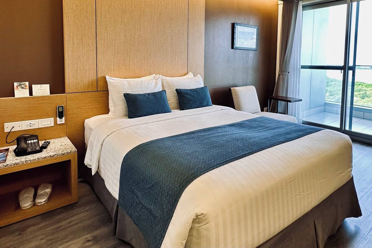 Habitación de hotel con una cama grande, una mesita de noche con teléfono y lámpara, una ventana con vistas a zonas verdes y un cabecero de madera.