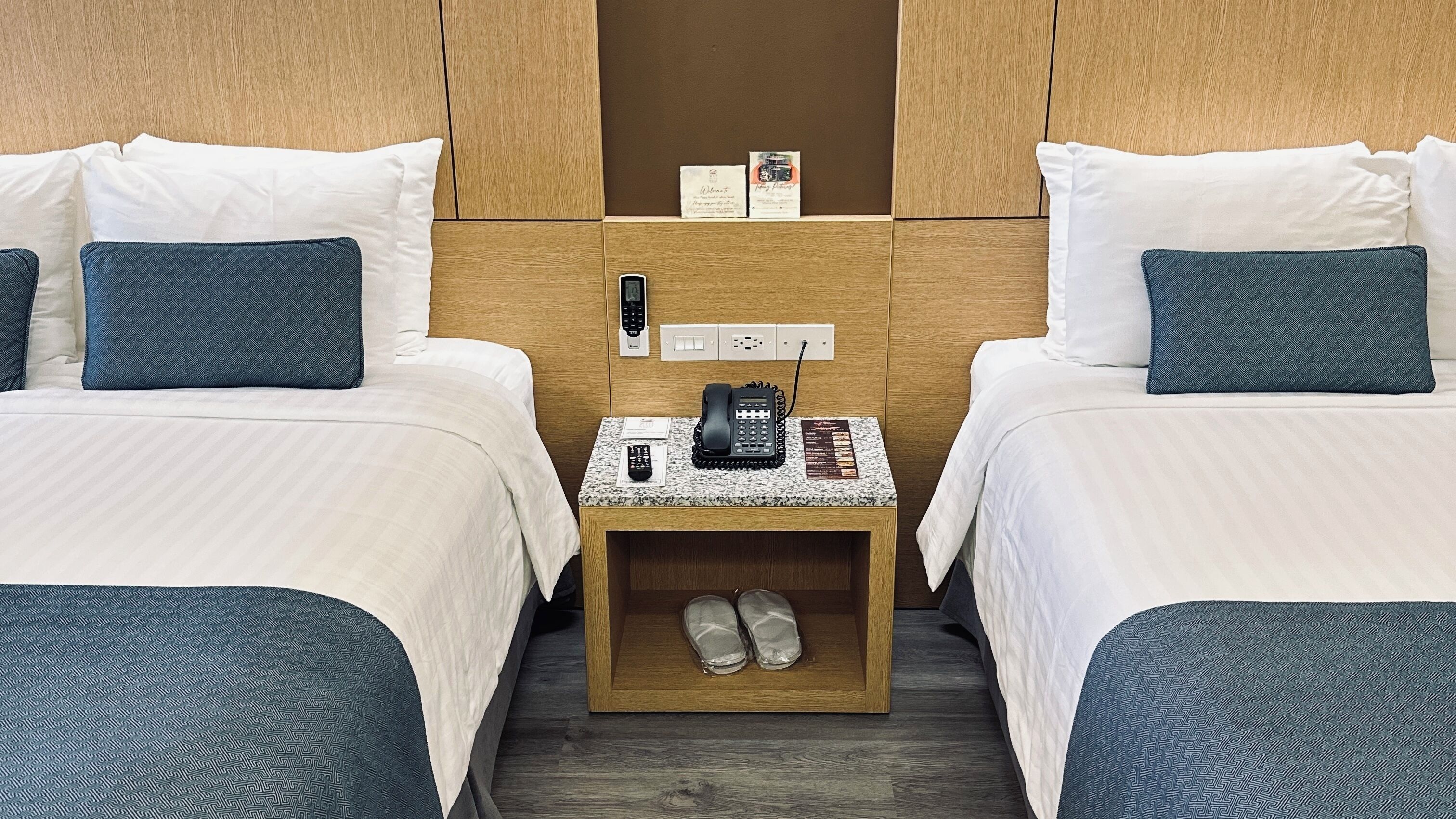 A hotel room with two beds, a small bedside table with a phone, a remote control, and slippers.