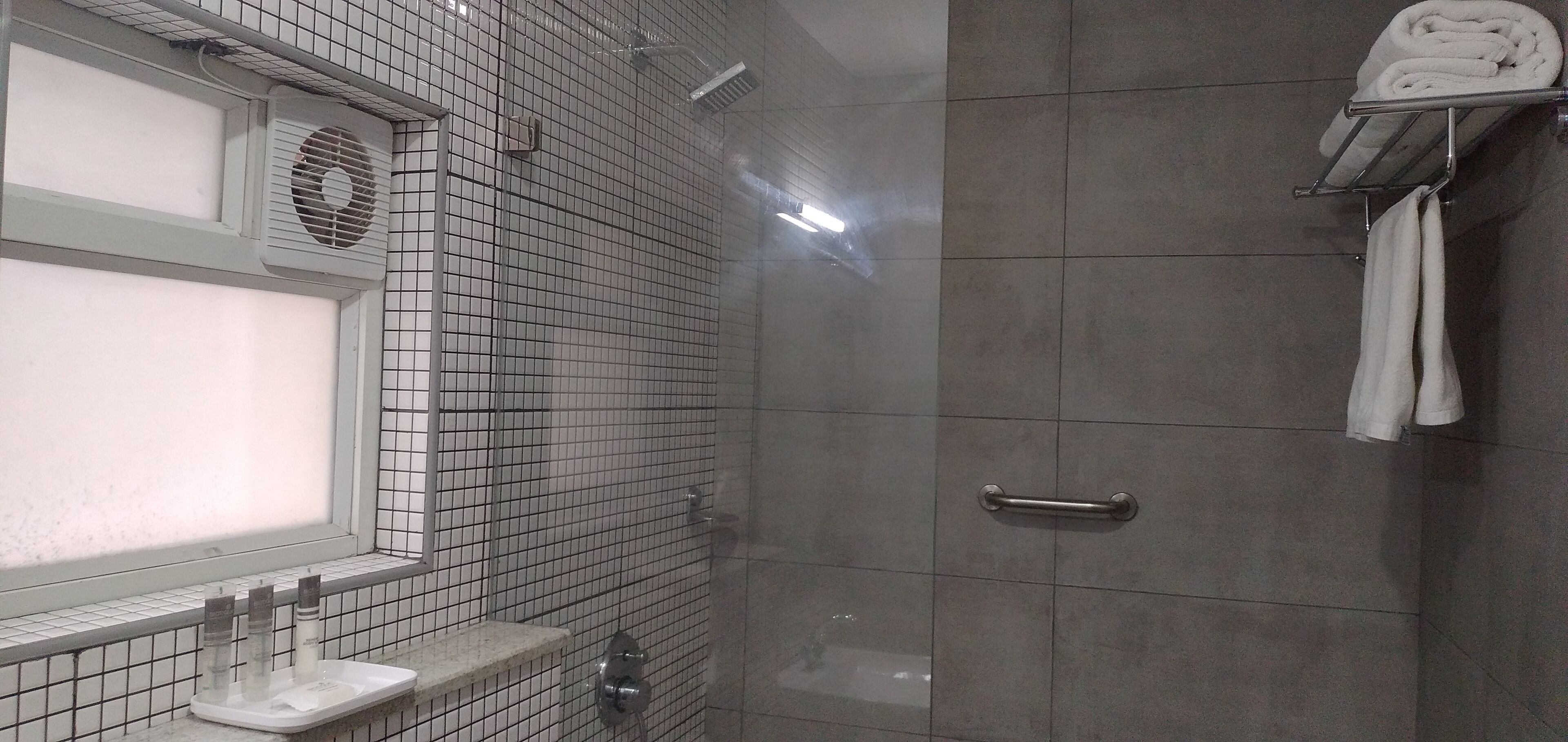 Ein modernes Badezimmer mit gefliesten Duschwänden, einem Fenster mit Jalousien, einem Handtuchhalter und einem Waschbecken mit Pflegeprodukten.