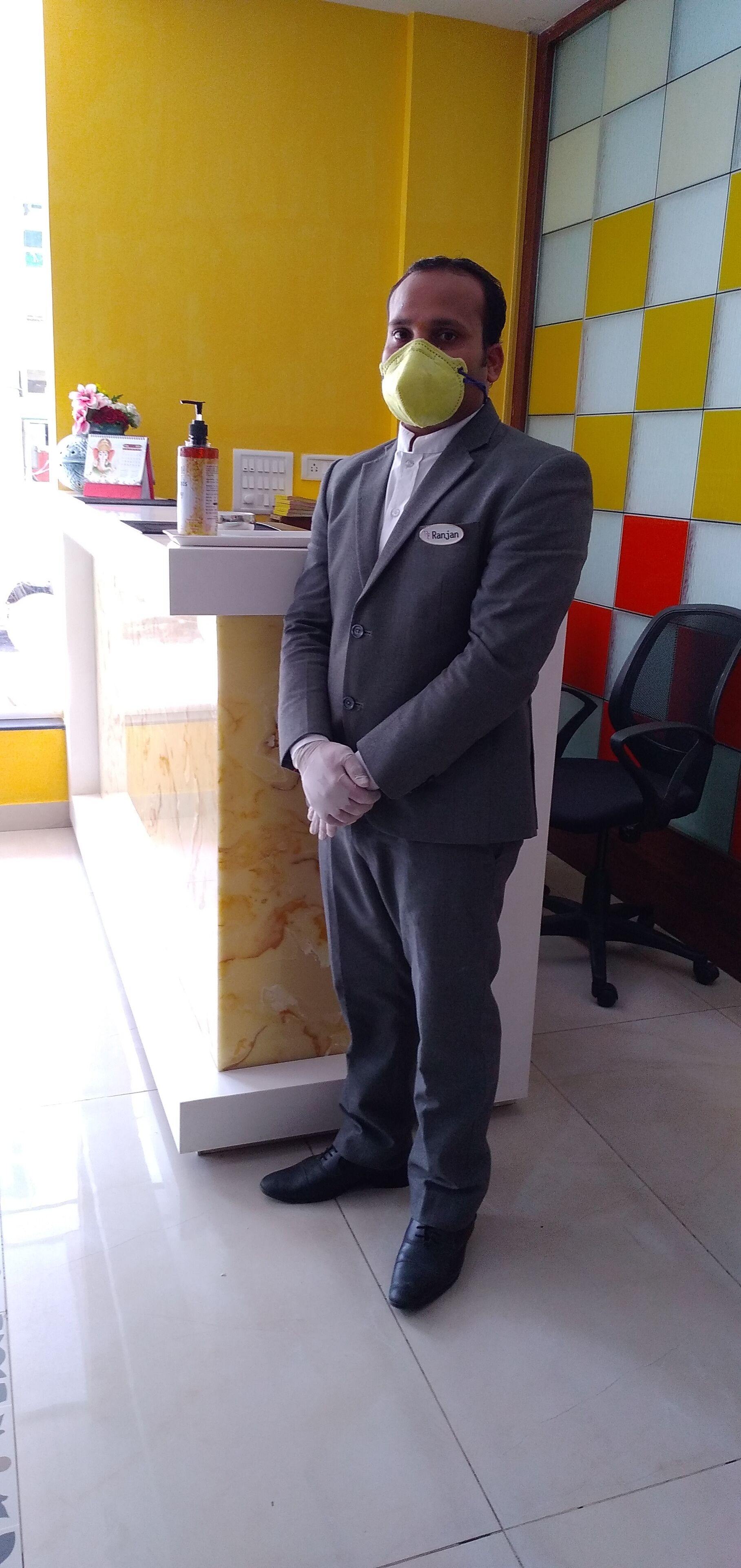 A man in a suit standing in a reception area with a marble counter, a name tag, and a yellow wall with a grid pattern.