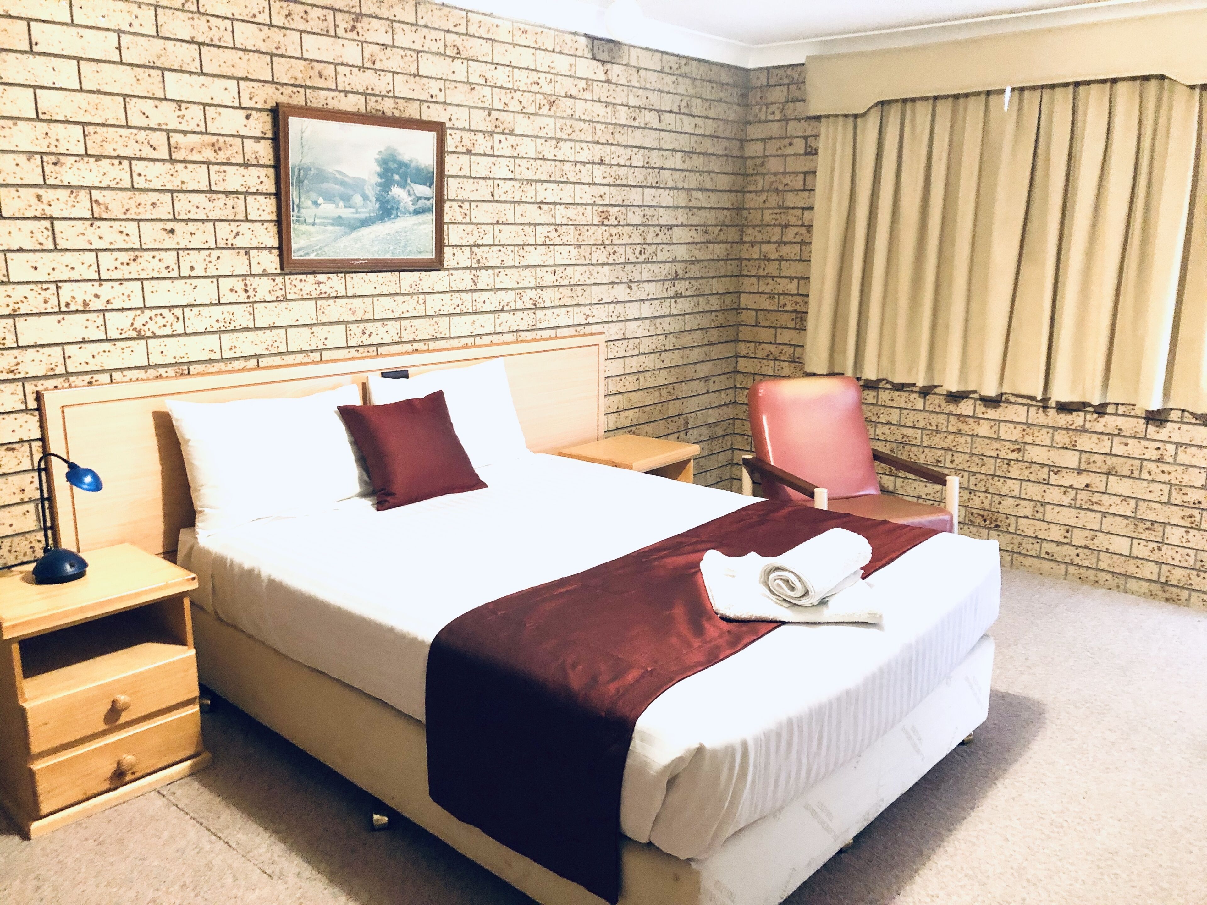 Una habitación de hotel con cama, mesita de noche, silla, un cuadro en la pared y ventana con cortinas.