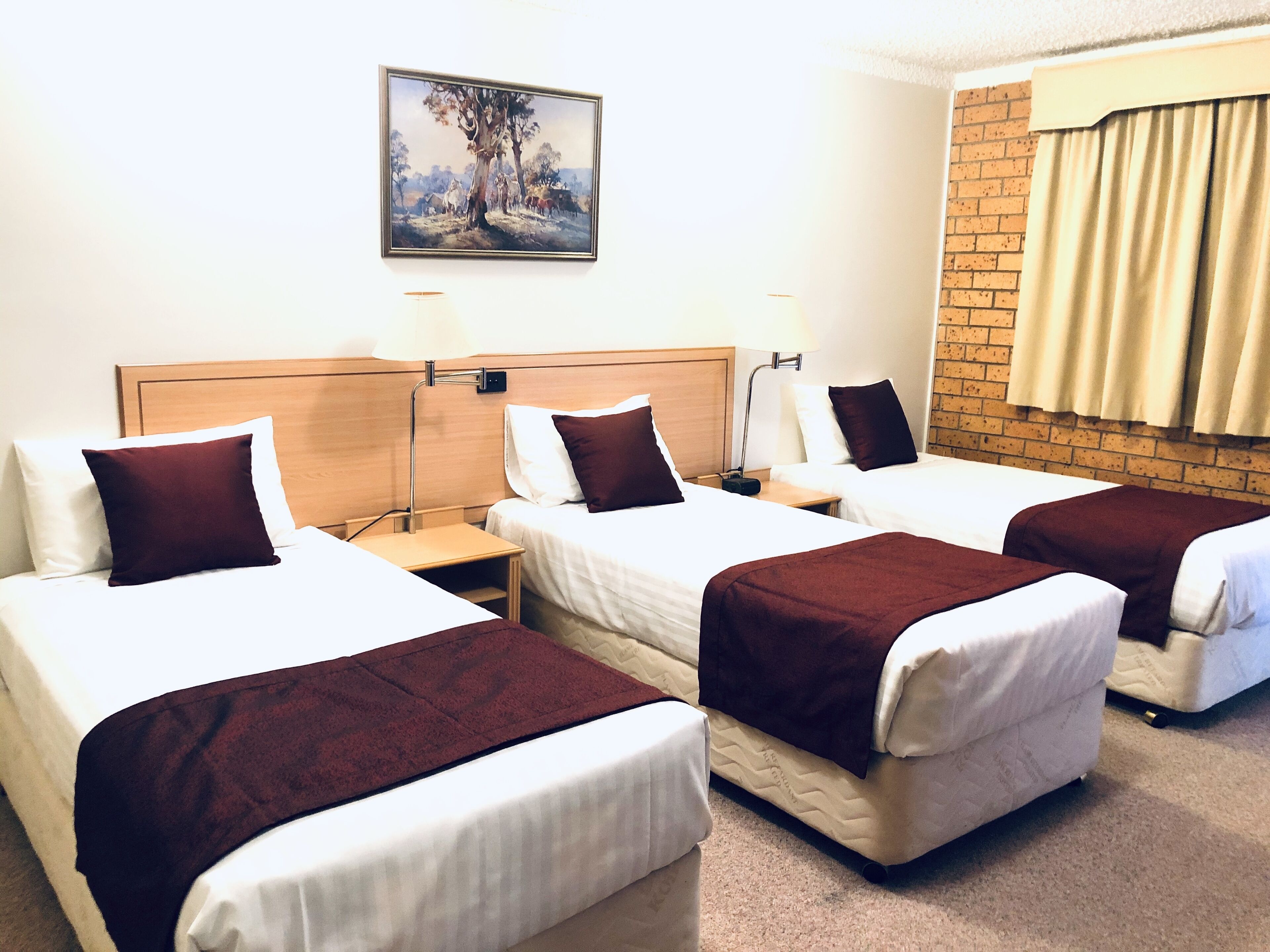 Une chambre d’hôtel avec trois lits simples, une tête de lit en bois, un tableau au mur et un mur en briques avec une fenêtre.
