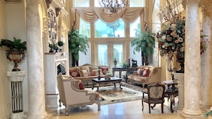 A luxurious living room with ornate furniture, large windows, and decorative plants.