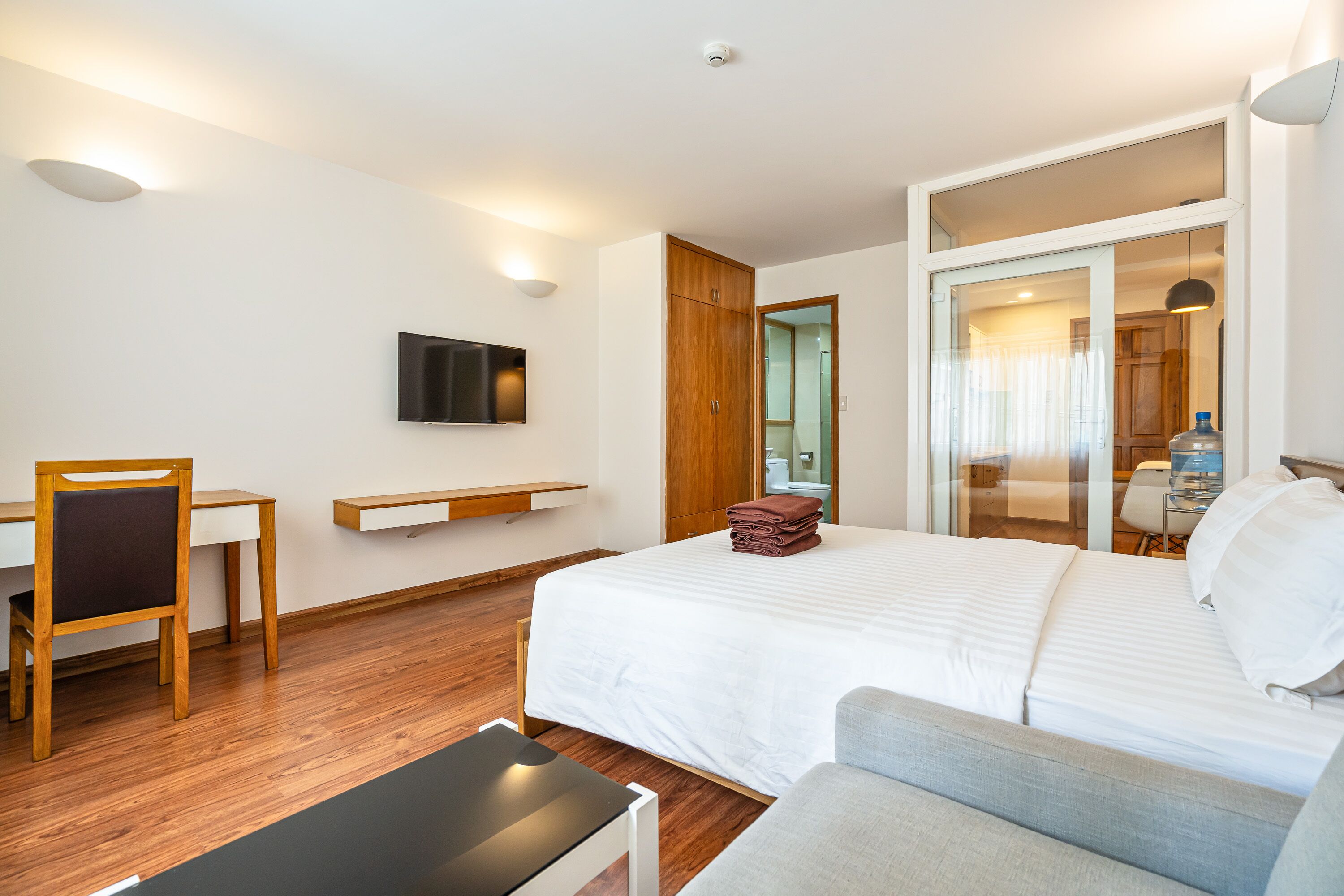 Ein modernes Hotelzimmer mit Bett, Schreibtisch, Stuhl, Wandfernseher und einer Glastür zum Badezimmer.