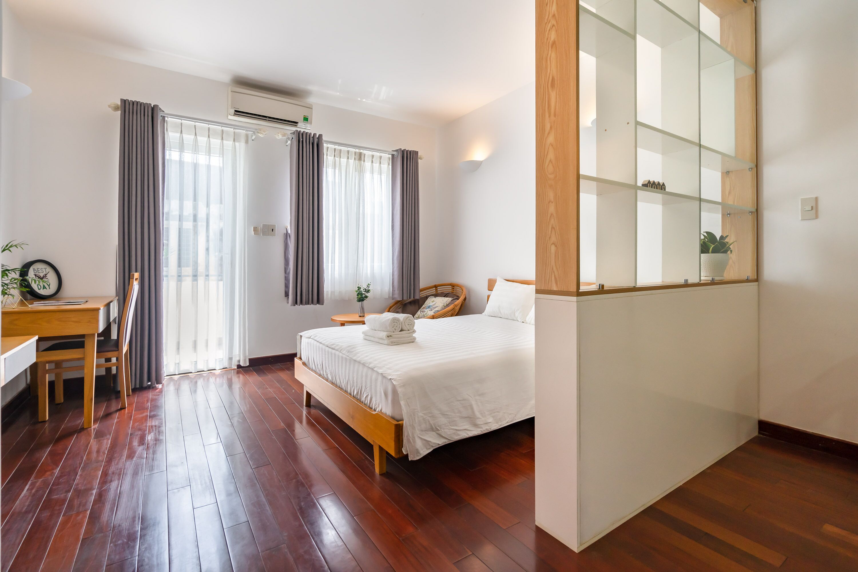 A bedroom with a wooden bed, a desk, and a chair. There is a window with curtains, a bedside table, and a mirror.