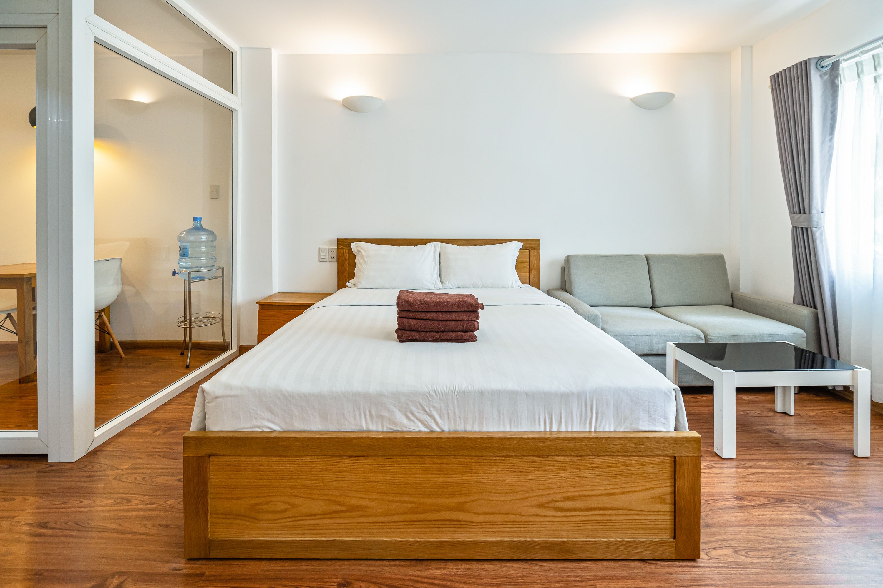 Ett modernt hotellrum med en säng i trä, en grå soffa, ett litet bord och en stor spegel.