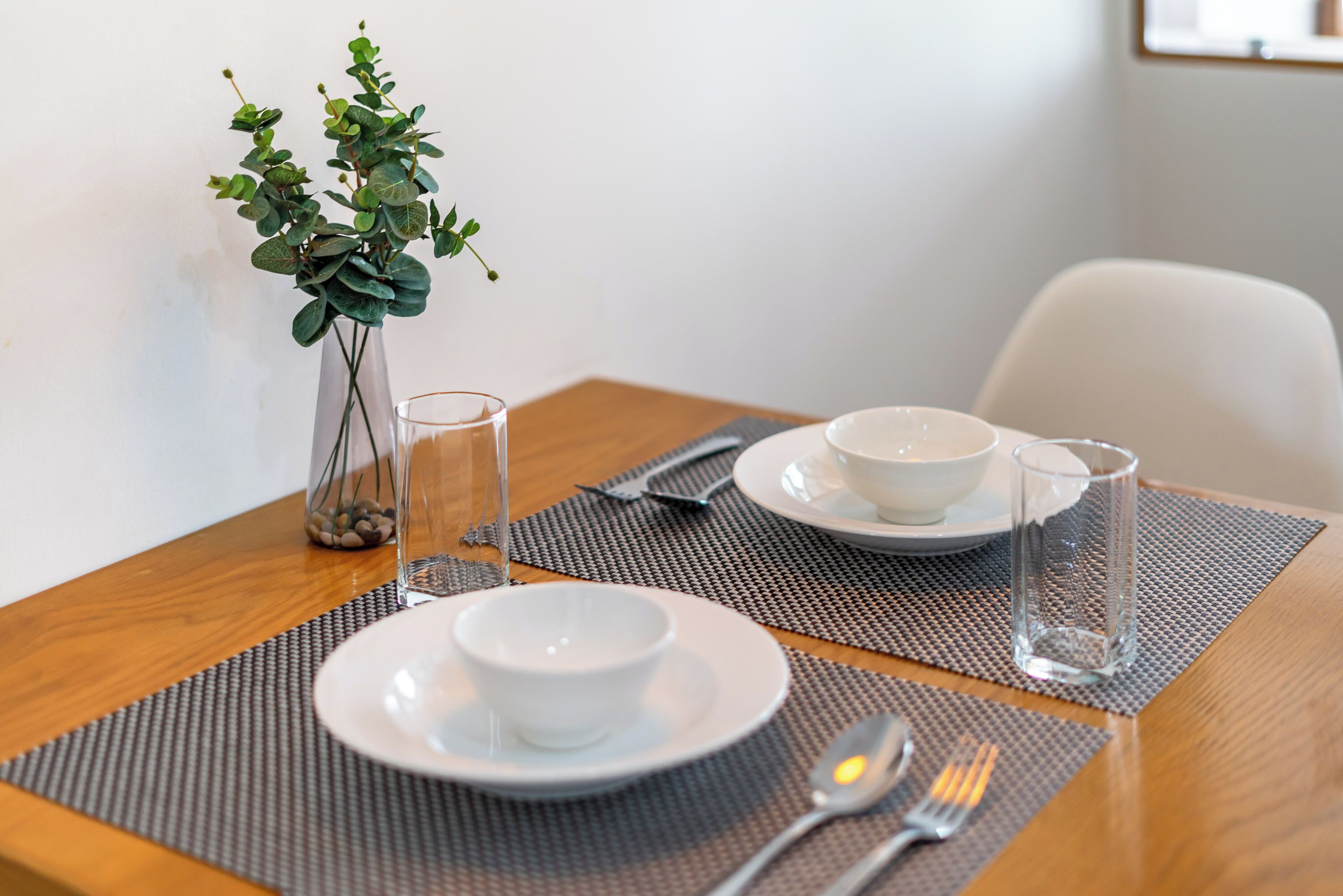A dining table with two white bowls, a glass, and a vase with green leaves.