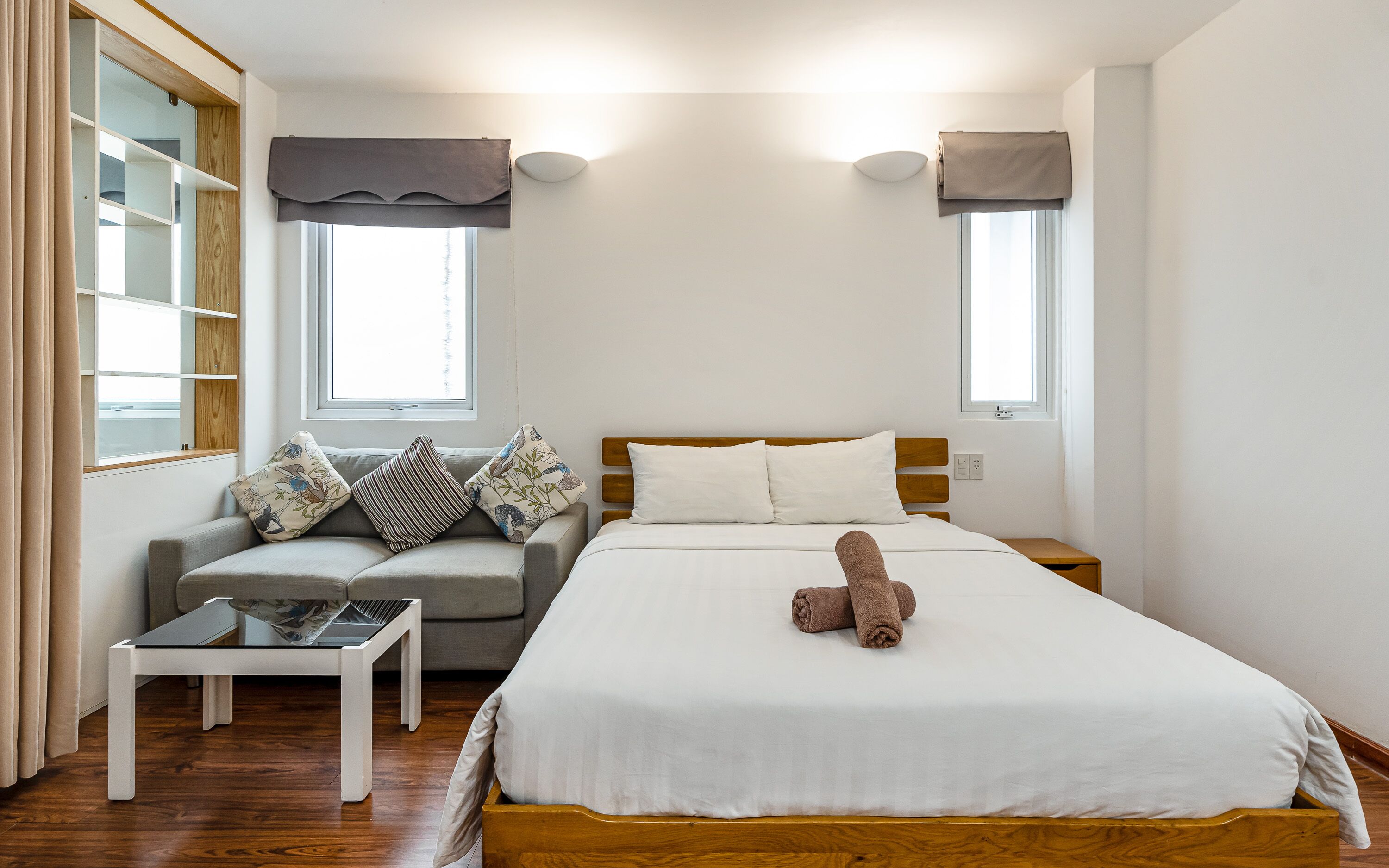 Ein Schlafzimmer mit Bett, Sofa, kleinem Tisch und zwei fensterbepflanzten Fenstern.