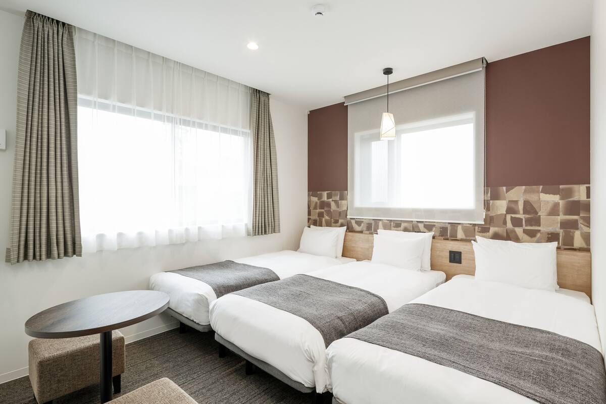 A hotel room with three beds, a small round table, and a window with curtains.