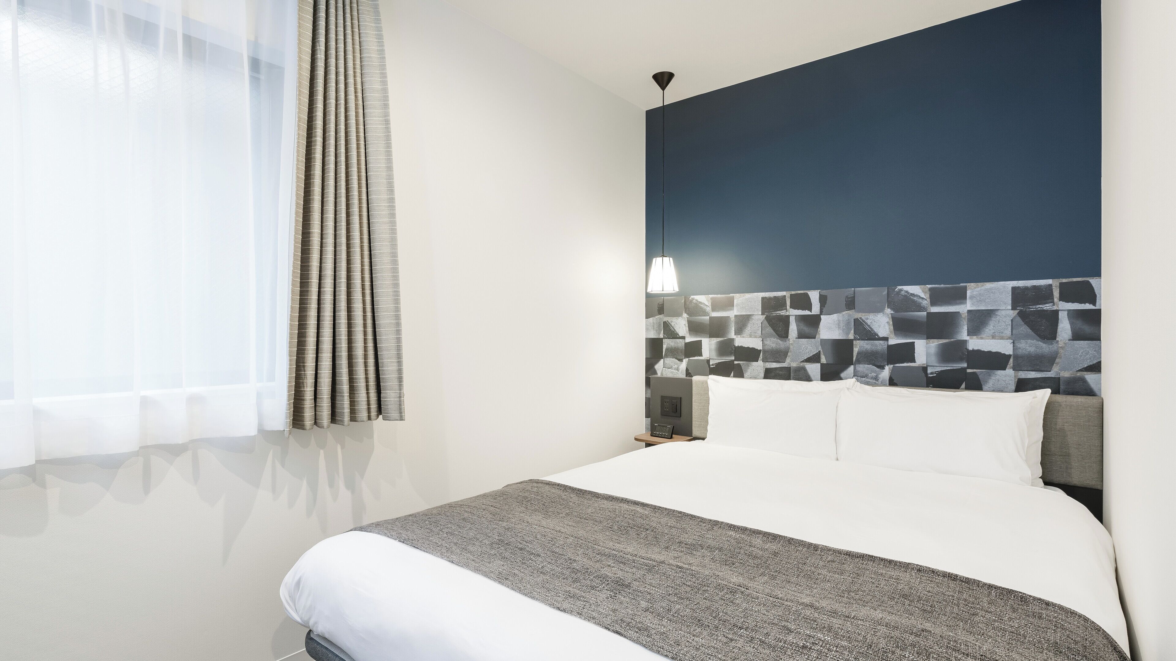 A hotel room with a bed, a nightstand, a wall-mounted light, and a window with curtains.