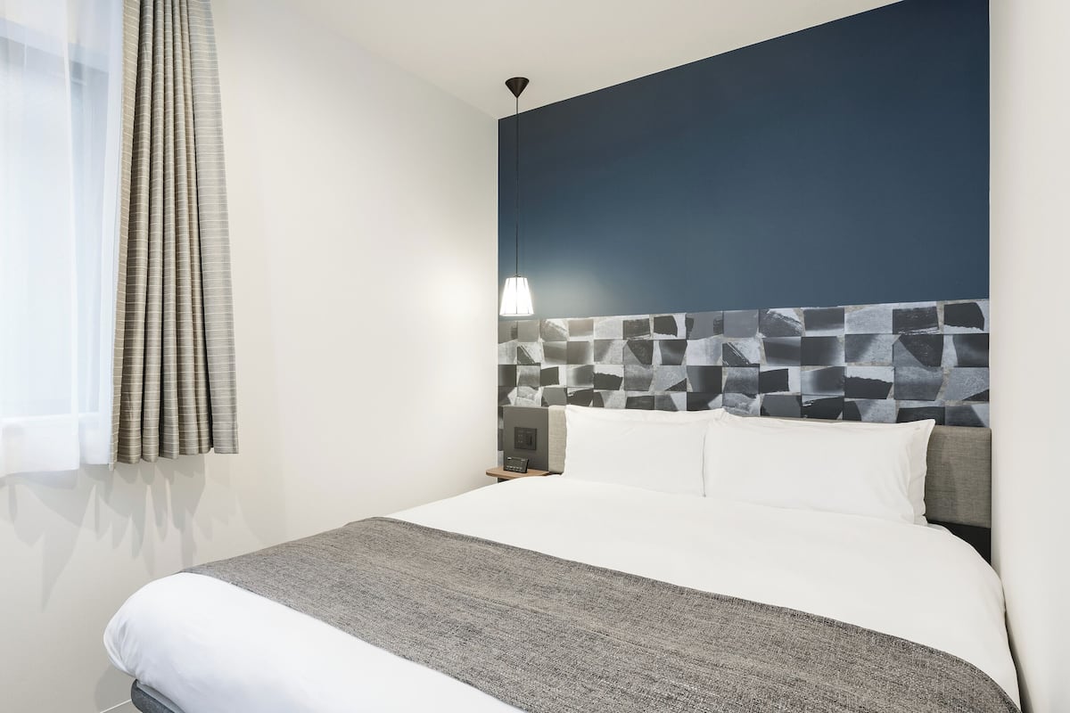 A hotel room with a bed, a nightstand, a wall-mounted light, and a window with curtains.