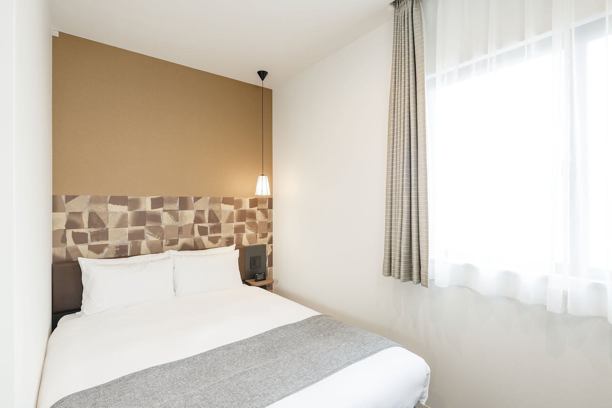 A neatly made bed with a grey throw blanket, a headboard with a mosaic pattern, a bedside lamp, and a window with sheer curtains.