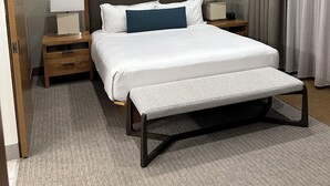 Une chambre d’hôtel comprenant un lit, des tables de chevet, un banc, une tête de lit et une fenêtre avec des rideaux.
