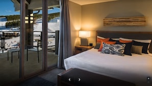 Habitación de hotel con cama, mesita de noche, lámpara y vista a un paisaje nevado a través de amplias ventanas.