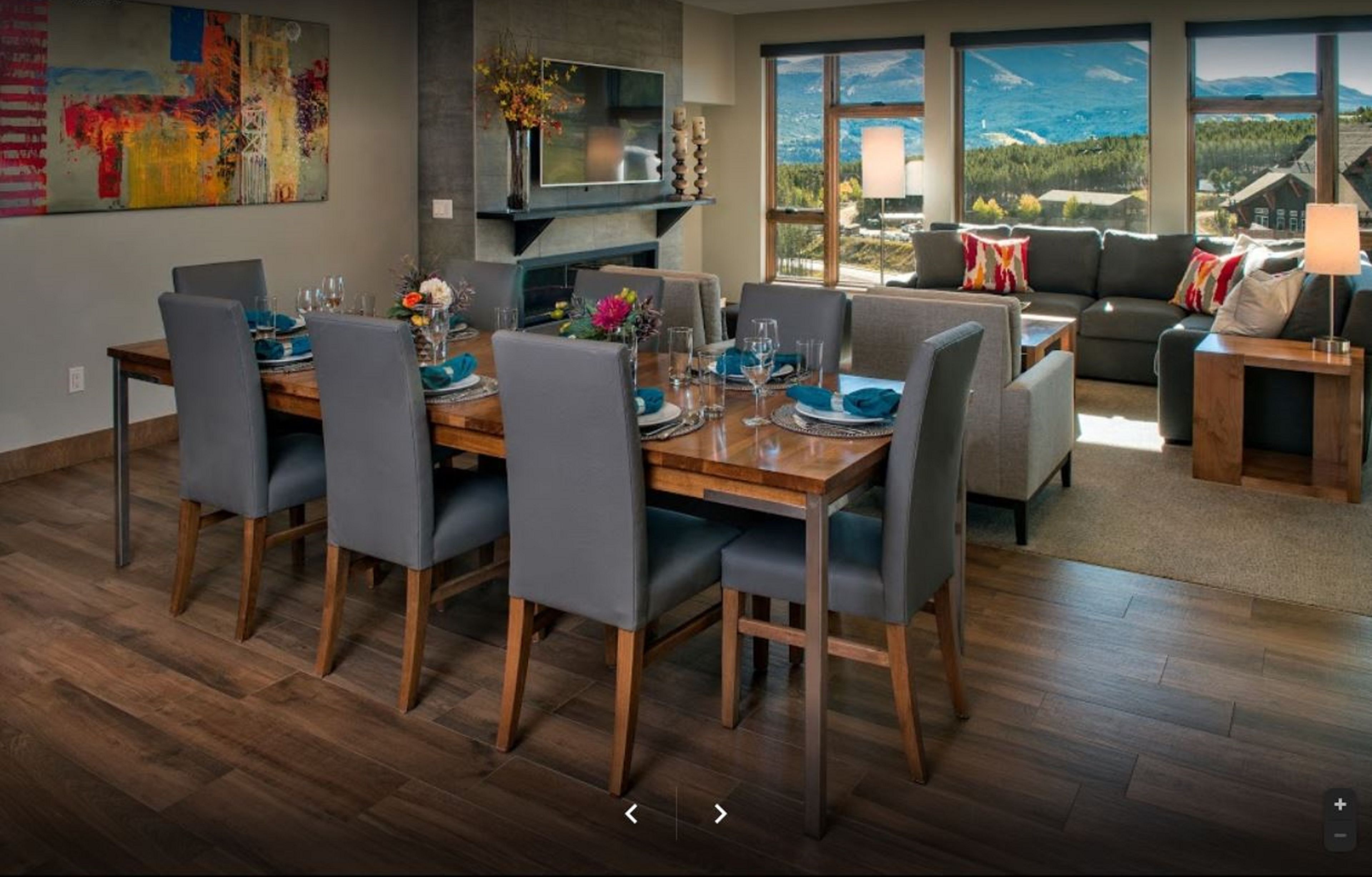 A modern dining area with a wooden table and grey upholstered chairs, a flat-screen TV mounted on the wall, and a large window offering a scenic mountain view.