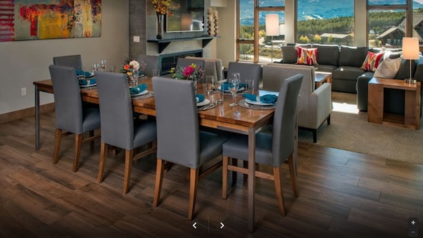 Un comedor moderno con una mesa de madera y sillas grises tapizadas, un televisor de pantalla plana montado en la pared y un ventanal con vistas panorámicas a la montaña.