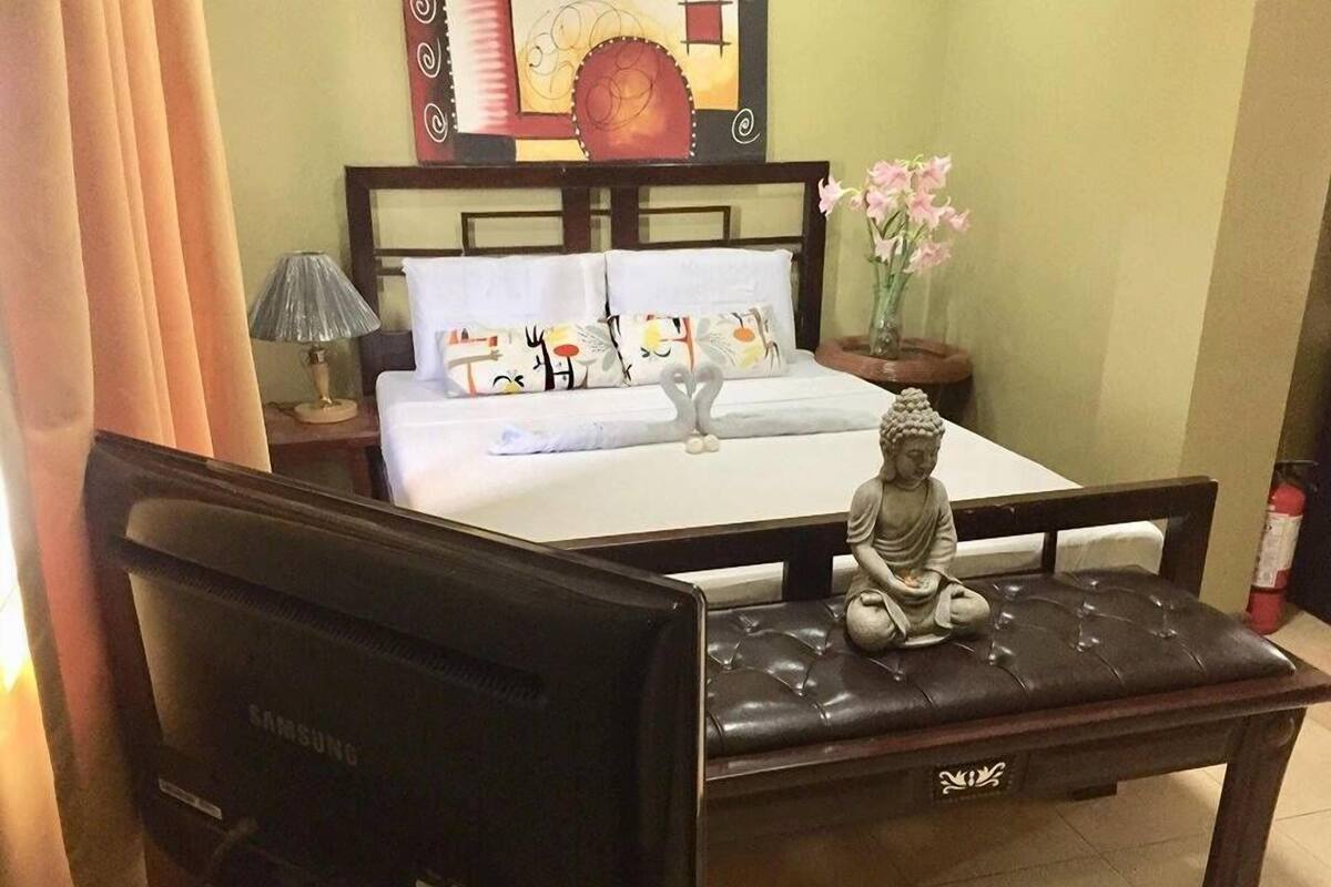 A hotel room with a bed, a bedside lamp, a Buddha statue, and a Samsung TV on a desk.