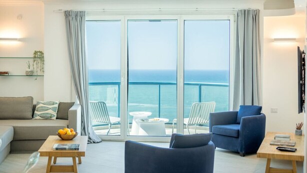 Un salon moderne avec un canapé, des fauteuils et une table basse. Une grande fenêtre offre une vue sur la mer.