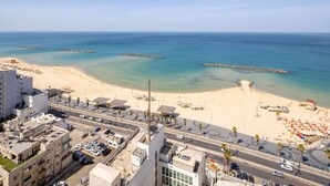 Un paysage urbain côtier avec une plage de sable, une mer bleue et limpide, et une route menant à un parking.